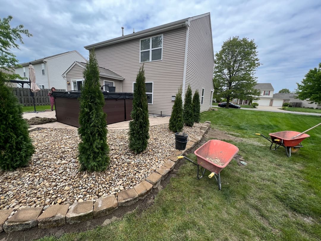 Backyard landscaping featuring stone pathways, shrubs, and wheelbarrows near a house.