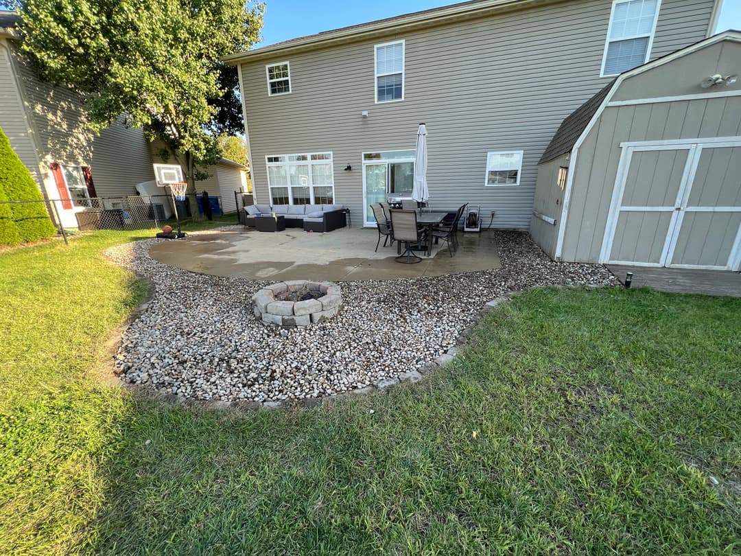 Backyard patio with seating area, fire pit, and shed, surrounded by decorative stones and grass.