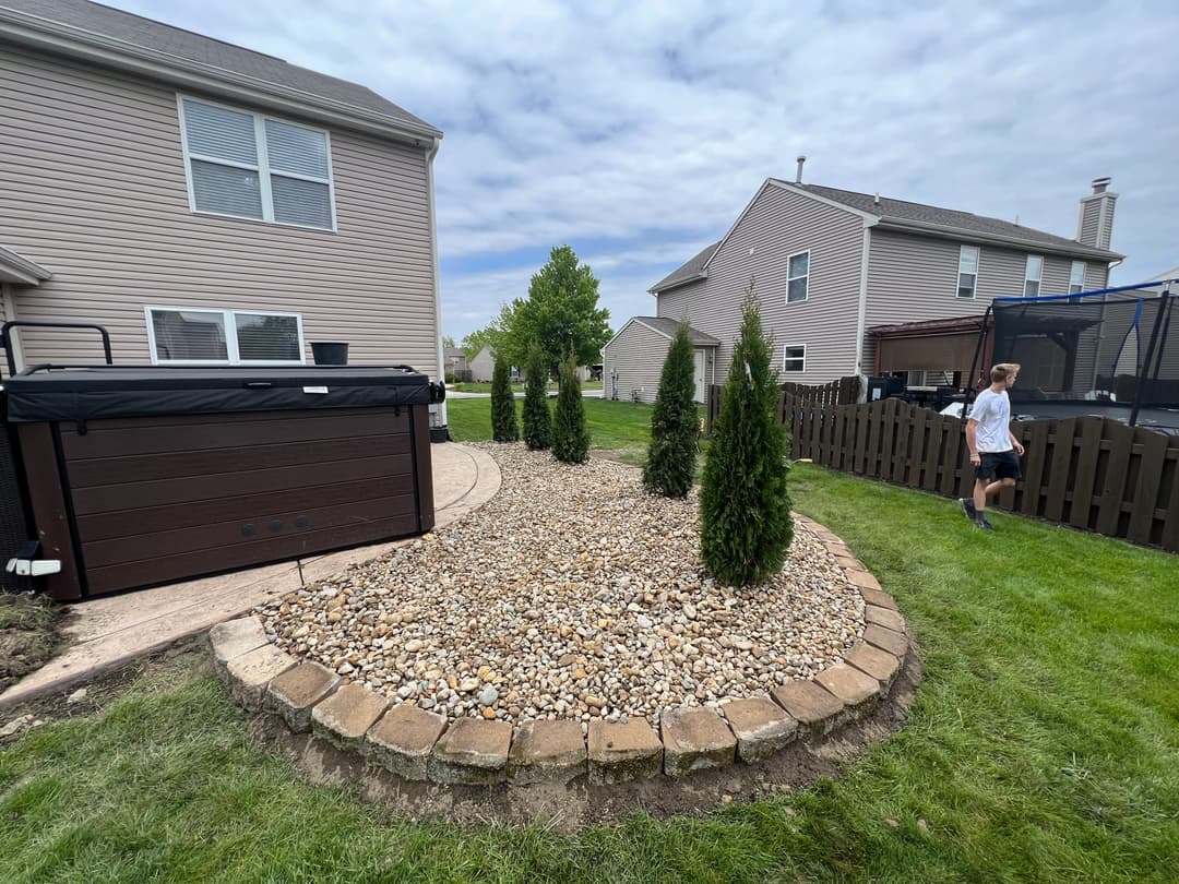 Backyard landscape featuring a gravel bed, evergreen trees, and a trampoline near a home.