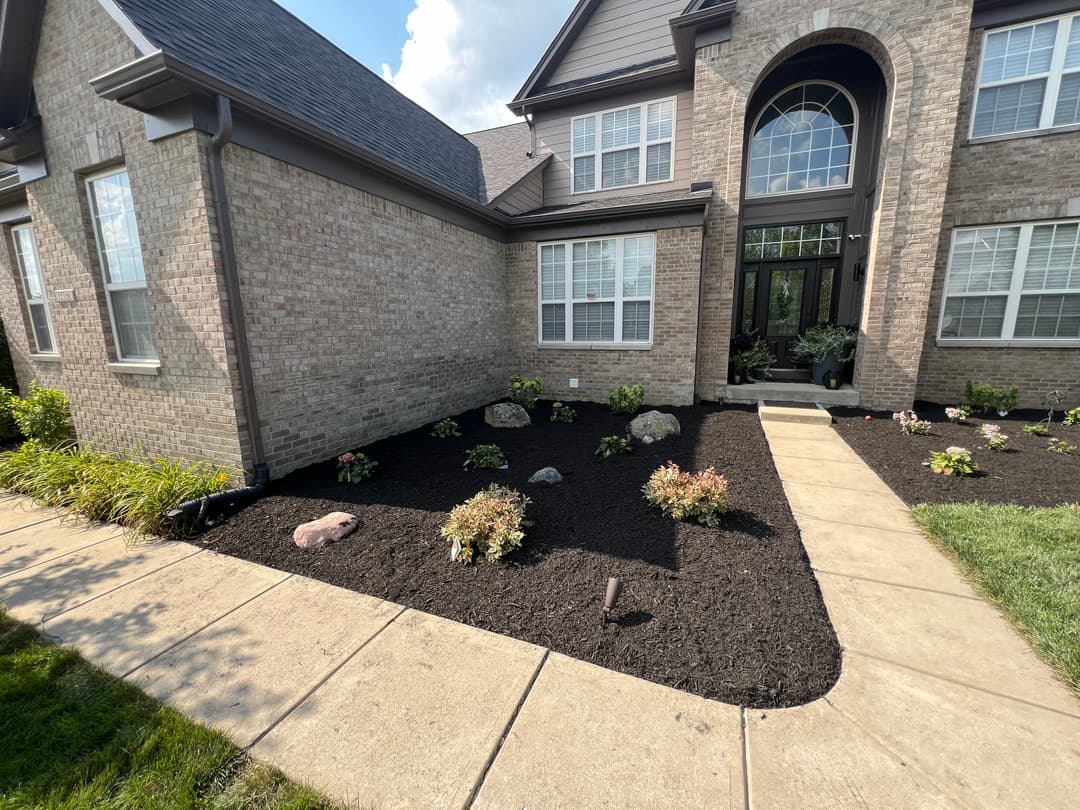 Front yard landscaping with mulched flower beds, plants, and a stone pathway leading to a house.