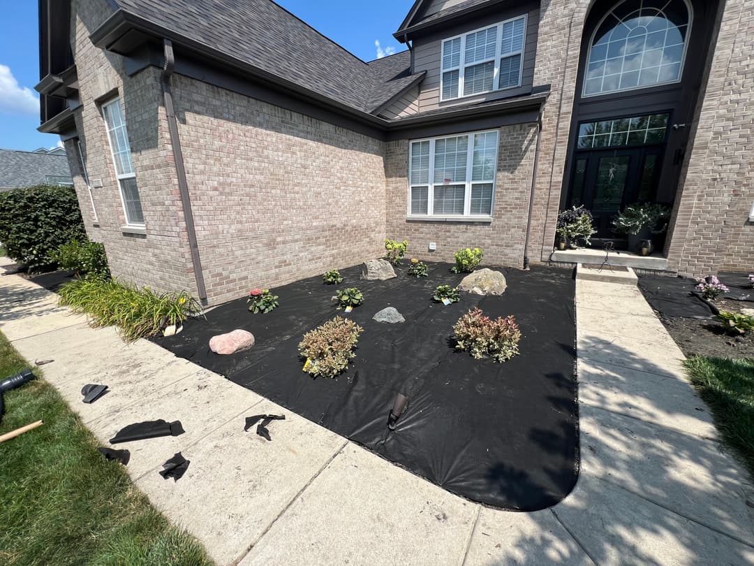 Landscaped front yard with rocks and plants on black fabric mulch beside a brick house.