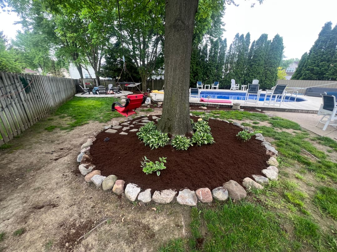 Mulched landscape bed around a tree with rocks and greenery, featuring a pool in the background.