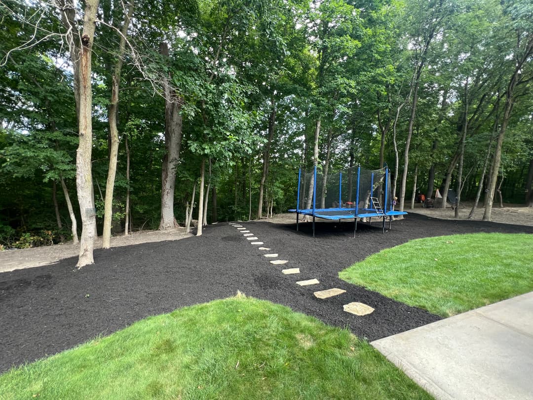 Trampoline in a wooded yard with stone path and freshly mulched landscaping.