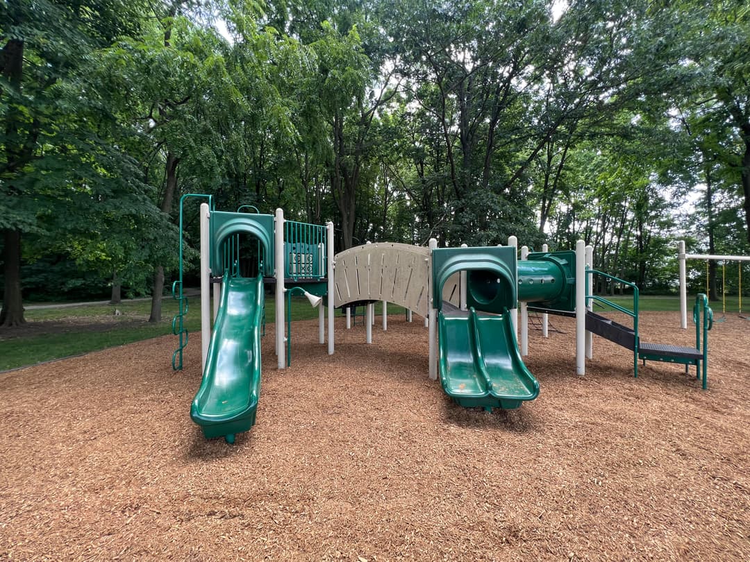 Green playground equipment with slides and a tunnel, surrounded by trees and wood chips.