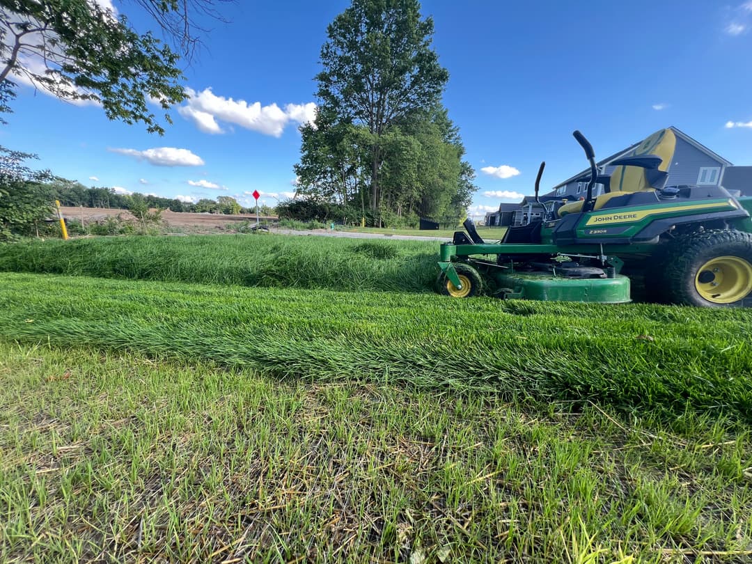 John Deere mower cutting grass in a lush green field under a blue sky with fluffy clouds.