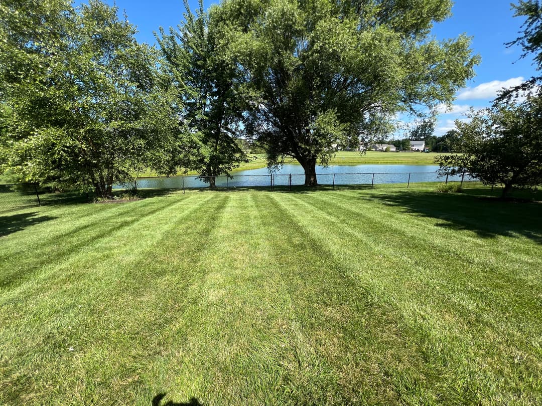 Lush green lawn with striped grass, trees, and a serene pond in the background.