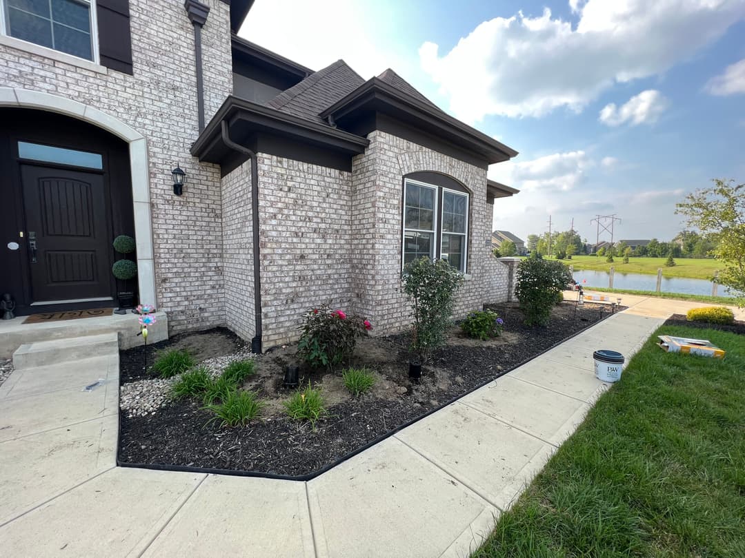 Landscape improvement of a home exterior with plants, flowers, and a sidewalk by a pond.