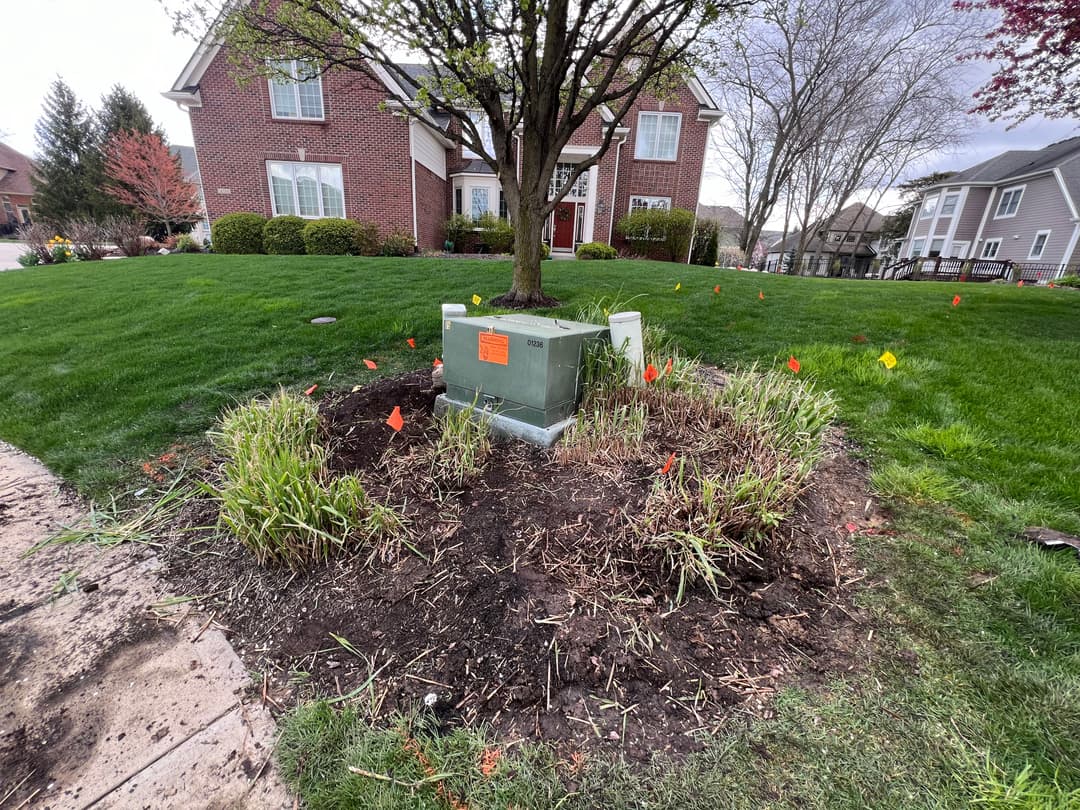 Utility box surrounded by grass, marked with flags, near a brick house and trees.