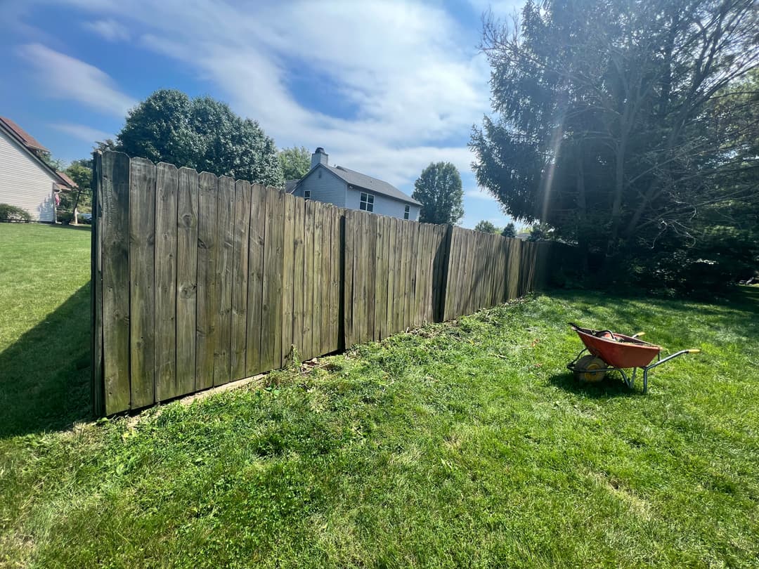 Wooden fence in a yard with a wheelbarrow on green grass and a house in the background.