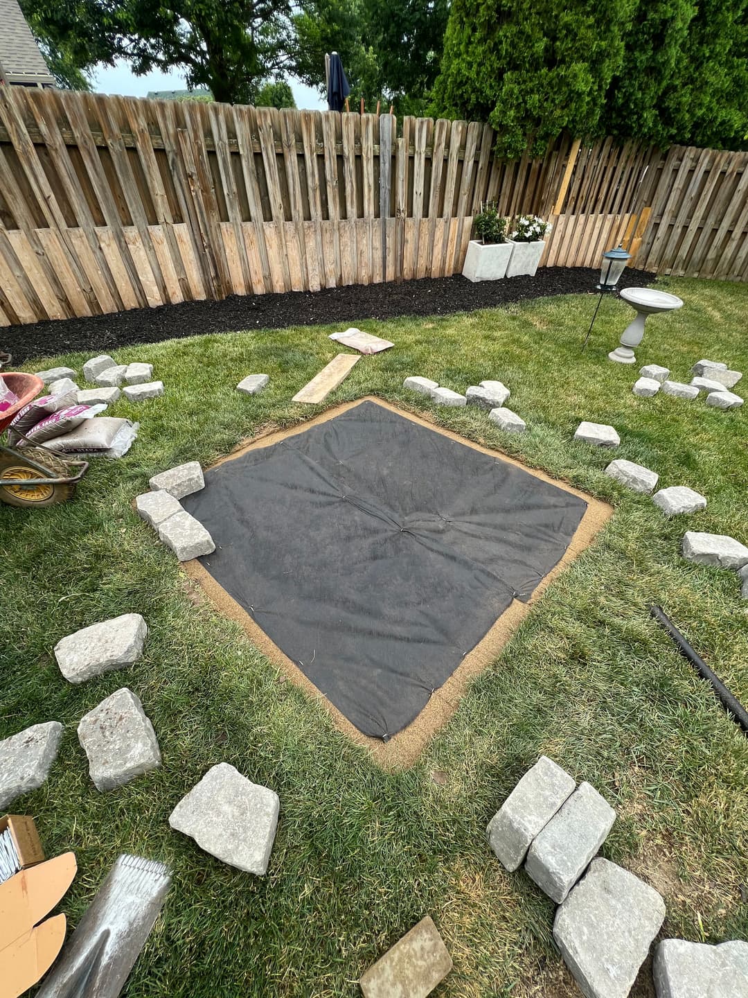 Grassy backyard with a prepared area for a square patio, surrounded by stones and tools.