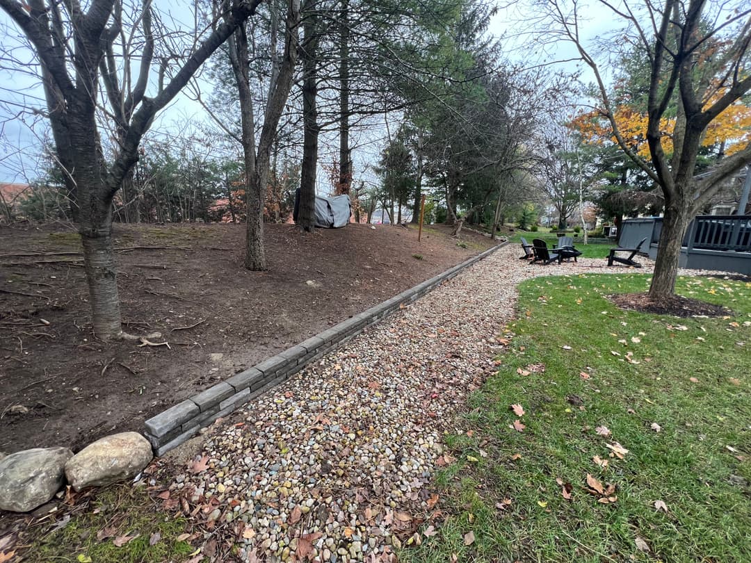 Gravel pathway lined with stones and trees, leading to a yard with outdoor seating.