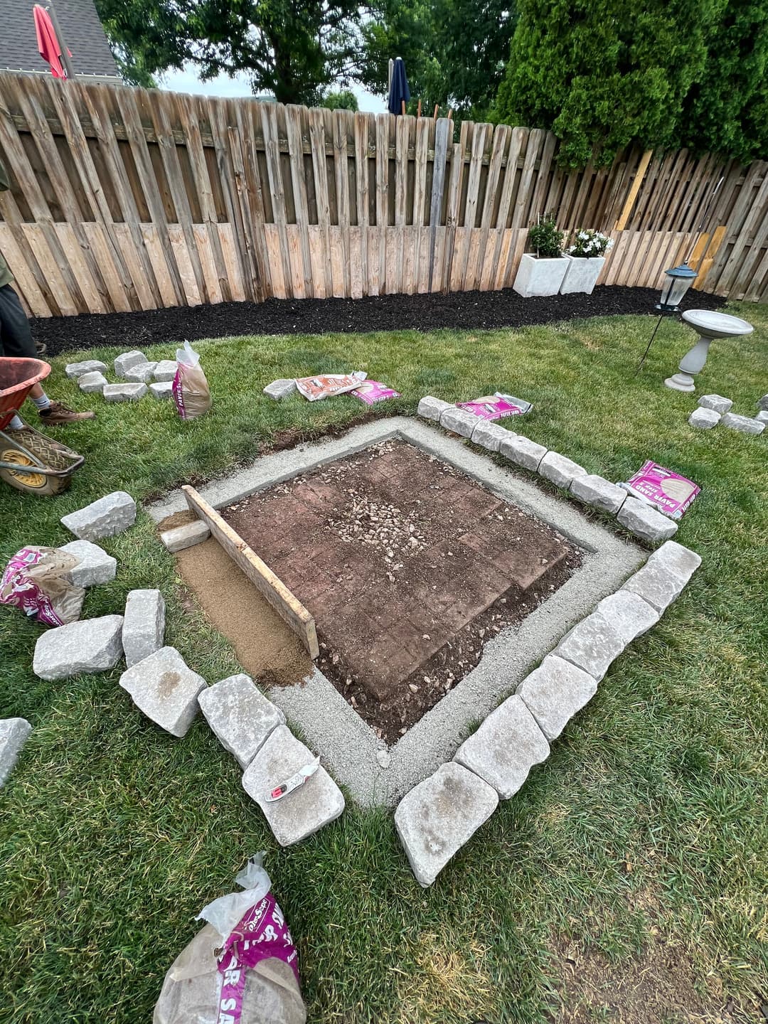 Square sandbox preparation in a backyard, surrounded by stones and landscaping materials.