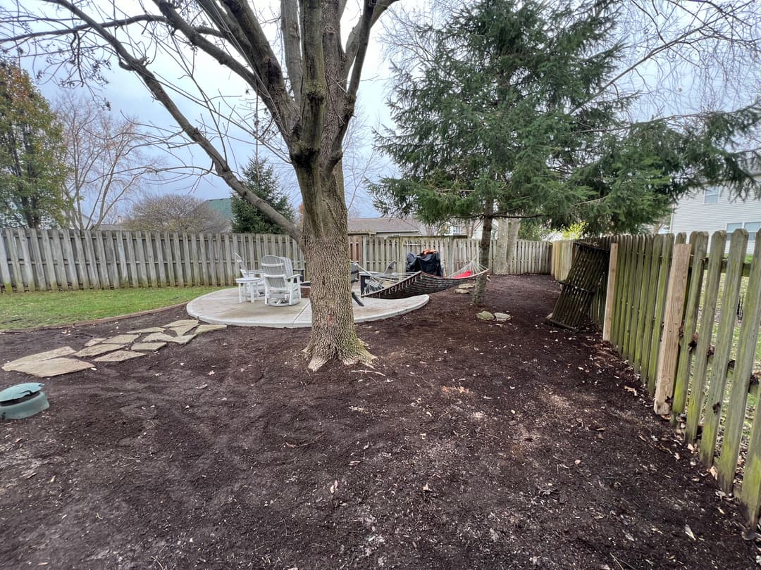 Backyard scene with a tree, hammock, and patio area, surrounded by a wooden fence.