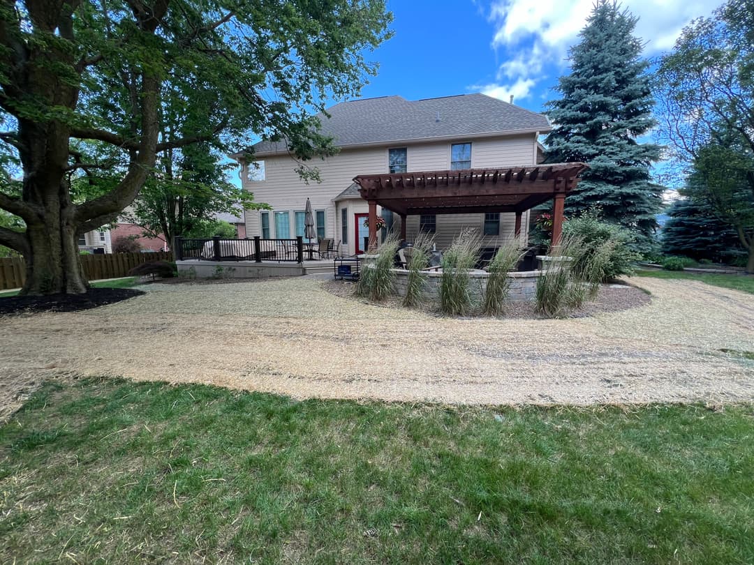 Backyard landscaping with a pergola, grass, and shrubs near a two-story house.