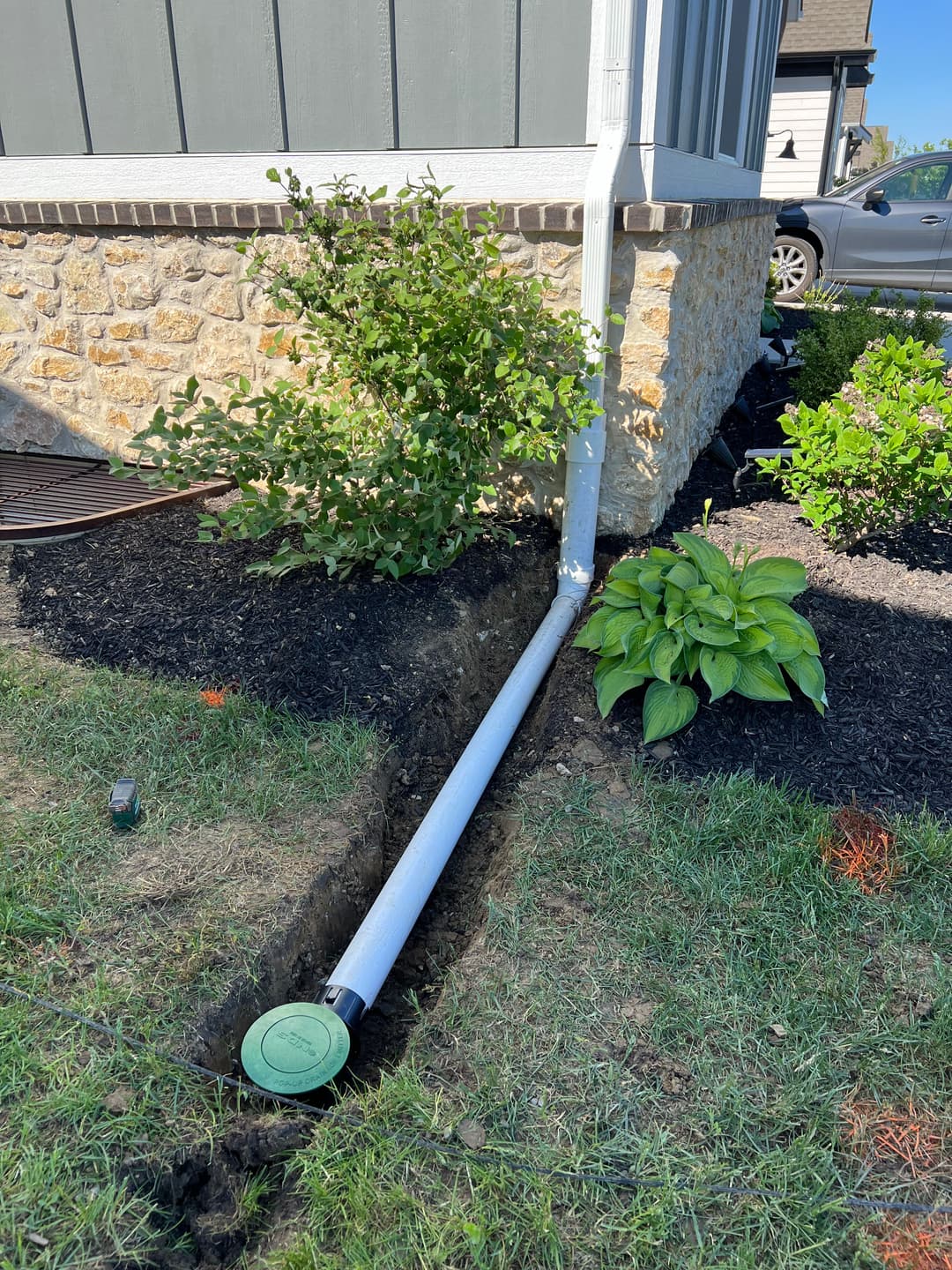 Drainage pipe installed along house foundation with grass and plants nearby.