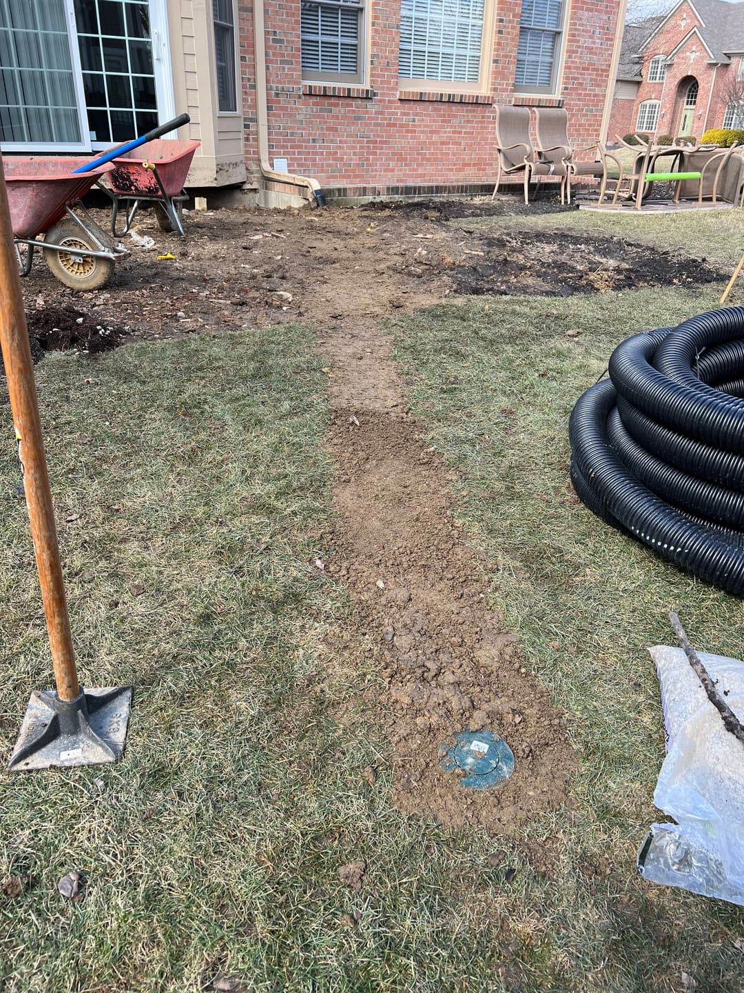 Dirt path in yard with wheelbarrow, shovel, and landscaping materials for drainage installation.