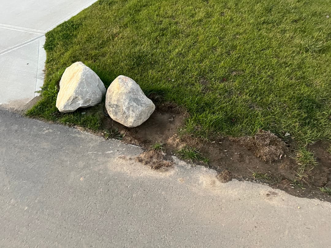 Two large rocks on grass beside a dirt patch near a concrete sidewalk.