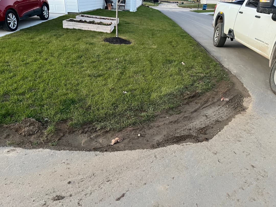 Dirt edge along a driveway with grass and a flower bed; vehicle parked nearby.