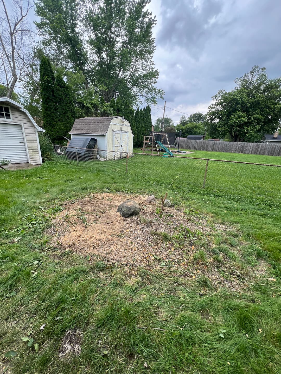 Garden area with a small dirt mound, shed, and playground equipment in a grassy yard.